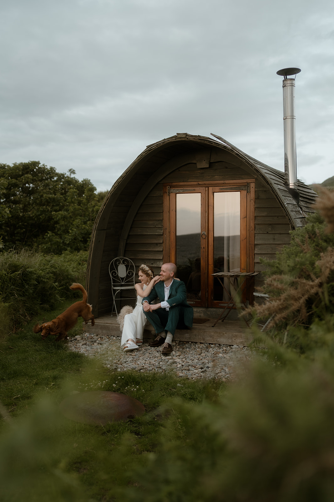 bride and groom sitting outside a cabin at sunset with their cute dog running free