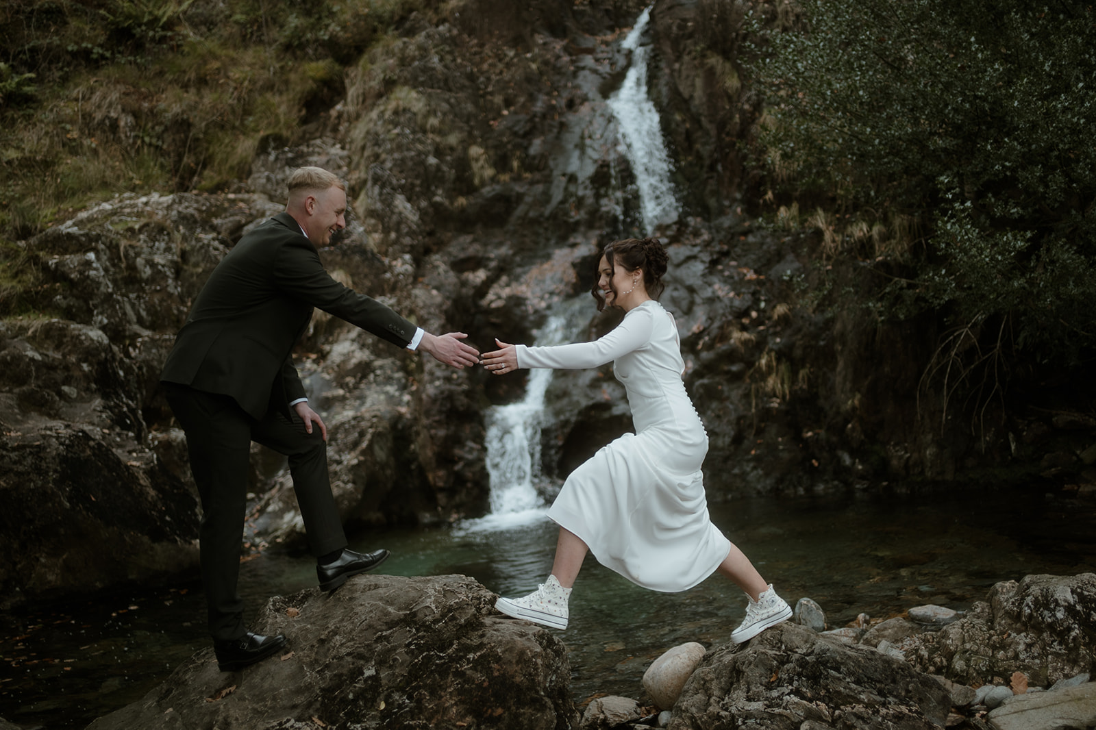 bride jumping across a stepping stone and being helped by the groom by a waterfall on their Elopement Day in Wales